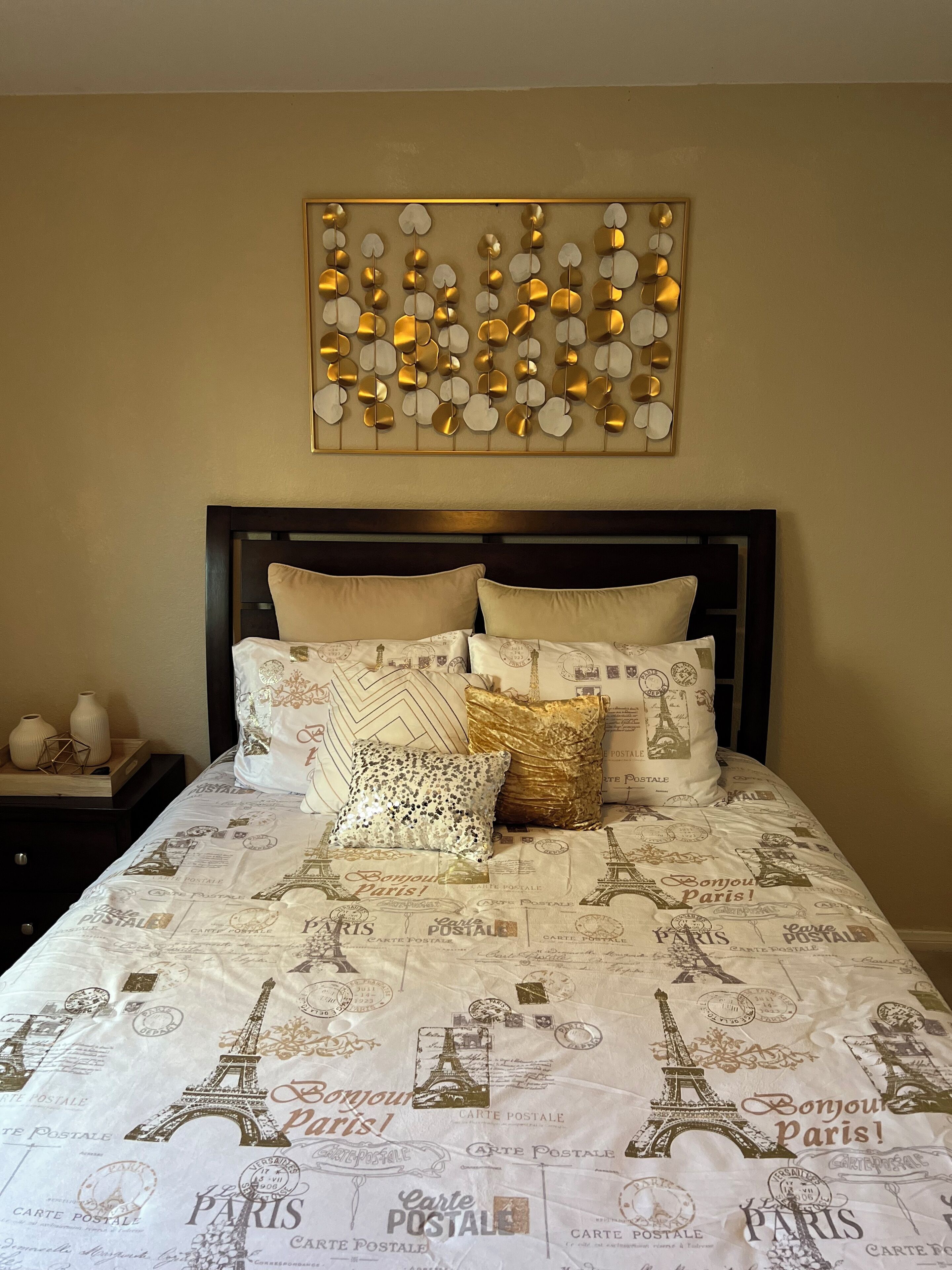 Un lit avec une couette aux motifs parisiens, un élément décoratif mural au-dessus, et une table de chevet agrémentée de vases.
