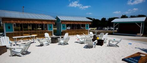 A beachside seating area with white Adirondack chairs, a swing set, and a thatched-roof building.