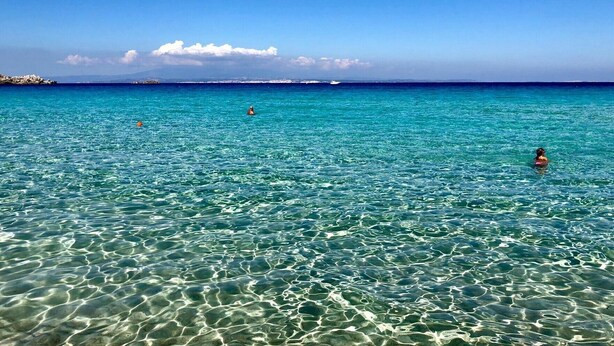A clear blue sea with visible underwater patterns, a distant rocky shoreline, and a clear sky.