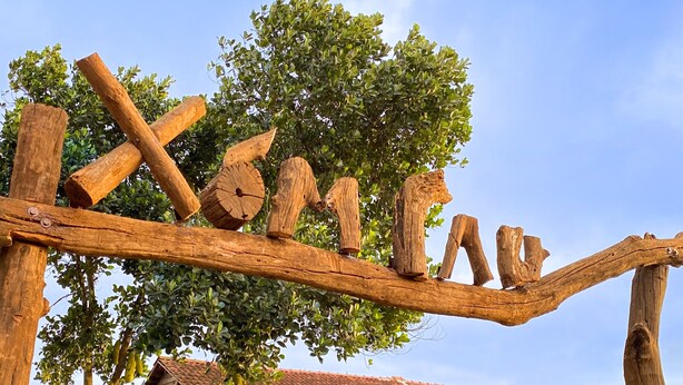 A rustic wooden sign with the text "XOMIA" in a natural setting, a house with a tiled roof, and a wooden fence.