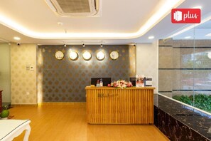 A reception area with a wooden reception desk, a wall clock, and a 'plus' sign indicating additional services. - Spring Airport Hotel (Ho Chi Minh City)