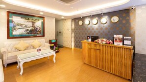 A reception area with a wooden reception desk, a white sofa with yellow cushions, and a painting of a tree on the wall. - Spring Airport Hotel (Ho Chi Minh City)