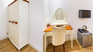 Una stanza con un armadio bianco, una scrivania in legno con uno specchio, un piccolo frigorifero e una televisione appesa al muro.