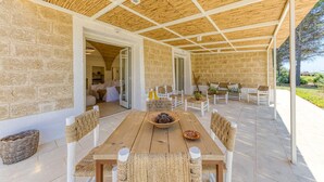 Un espace extérieur abrité, aménagé pour les repas, avec une table et des chaises en bois, des murs en pierre et une vue sur le jardin.