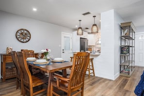 A dining area with a wooden table and chairs, a clock on the wall, pendant lights, and a bookshelf with books.