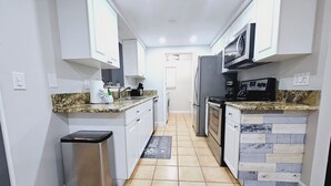 A narrow kitchen with white cabinets, a stainless steel refrigerator, a microwave, and a tiled floor. - 3BR retreat w/ private fence yard • 16 min to Siesta Key (Sarasota)
