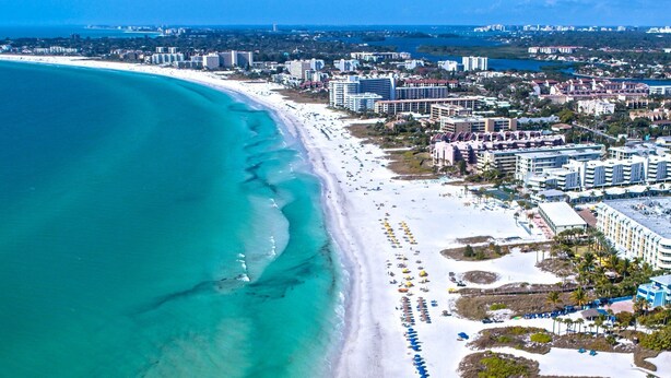 An aerial view of a coastal city with a wide sandy beach, adjacent high-rise buildings, and a clear blue sea. - 3BR retreat w/ private fence yard • 16 min to Siesta Key (Sarasota)