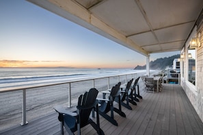 A covered deck with wooden chairs and tables, overlooking a beach with waves and a sunset.