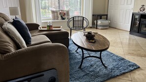 Een woonkamer met een bruine bank, een blauw vloerkleed, een bijzettafeltje, een rieten fauteuil en een open haard.