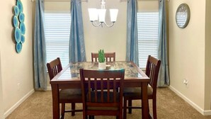Une salle à manger avec une table rectangulaire, six chaises en bois, un lustre et deux fenêtres ornées de rideaux bleus.