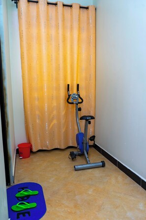 A small room with a standing exercise bike, a blue mat with "HOME" written on it, and a red bucket. - MLIMWA HILL PALACE (Dodoma)