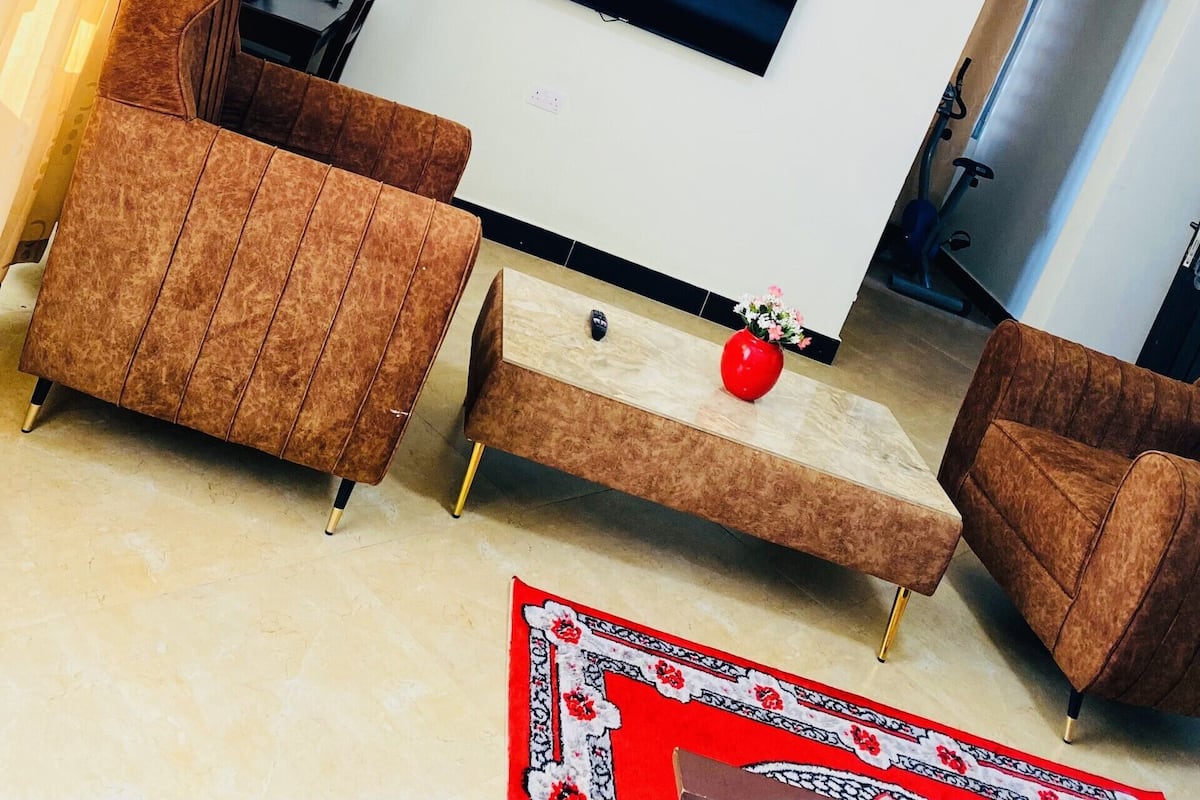 A living room with a flat-screen TV mounted on the wall, a brown sofa, a small coffee table, and a red vase.