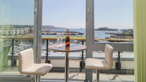 A balcony with two white chairs, a round table, and a bottle of wine. The view includes a marina, boats, and a distant shoreline. - The Penthouse, Phoenix Quay (Plymouth)