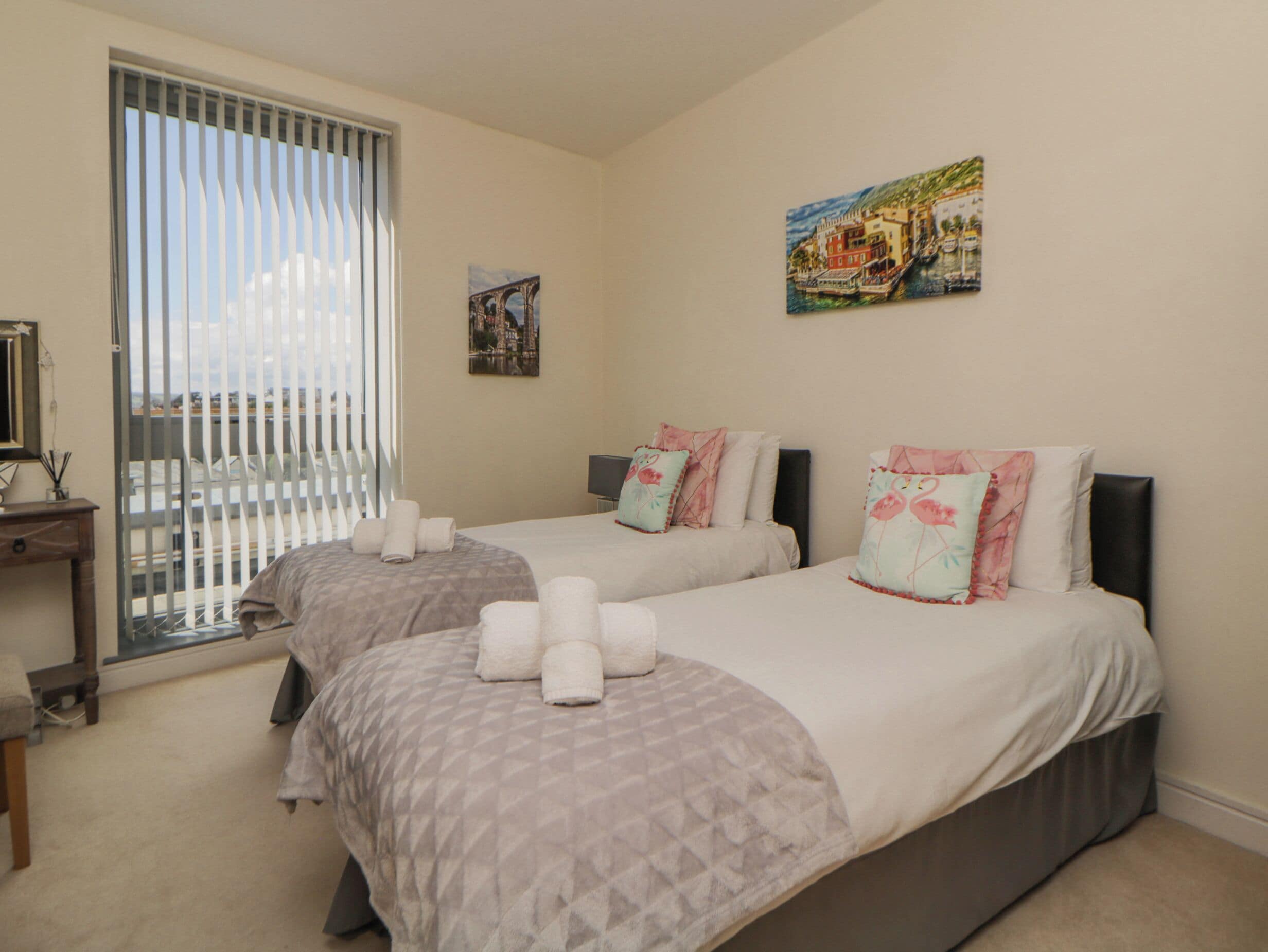 A hotel room with two beds, a desk with a chair, a window with blinds, and a painting on the wall.