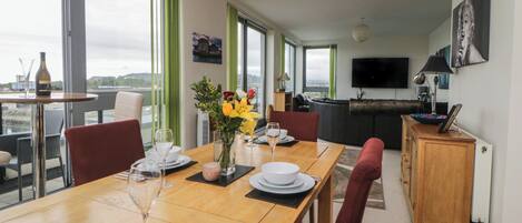A dining area with a wooden table set for four, a vase with flowers, and a view of the city through large windows.