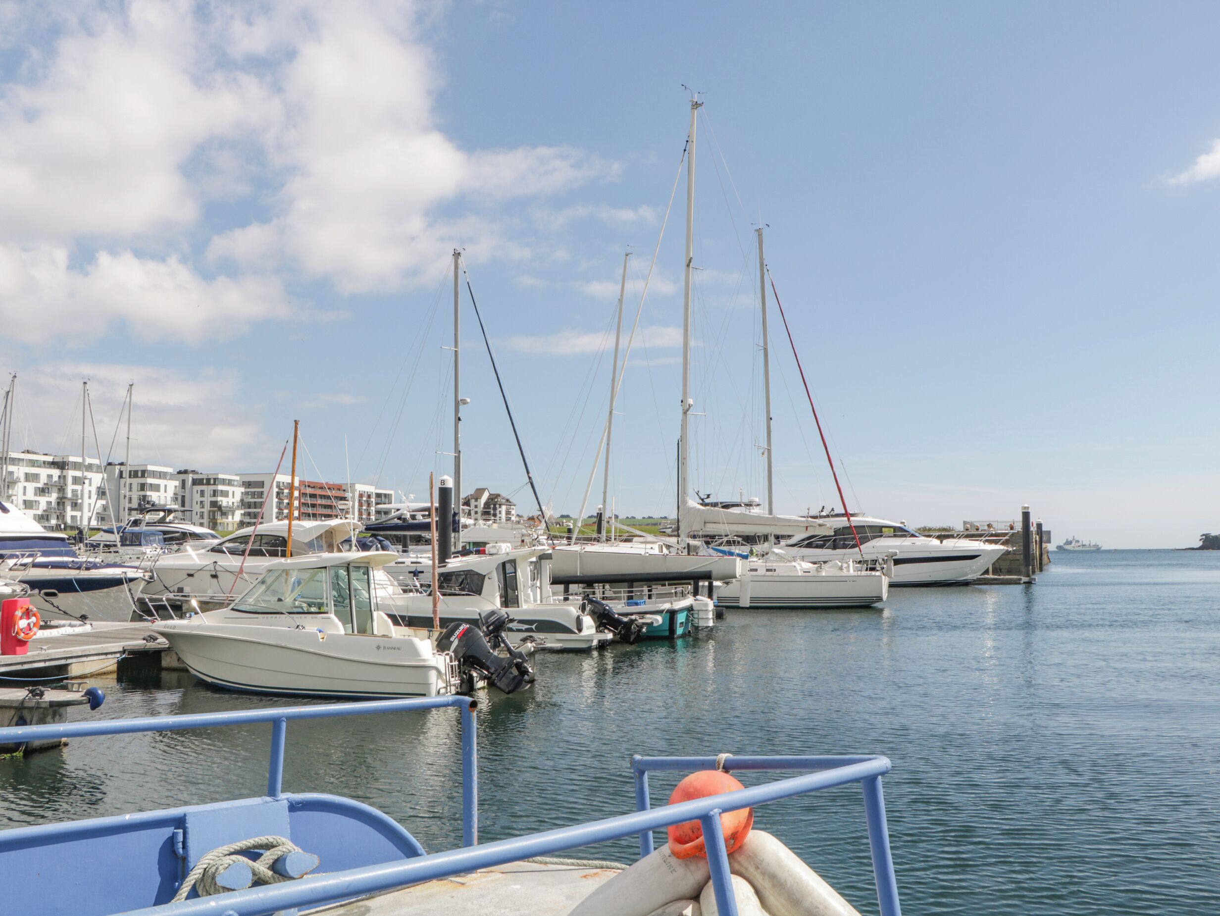 A marina with various boats docked, a clear sky, and buildings in the background.