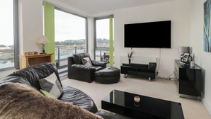 A modern living room with a large window, a black leather sofa, a black coffee table, and a flat-screen TV mounted on the wall. - The Penthouse, Phoenix Quay (Plymouth)