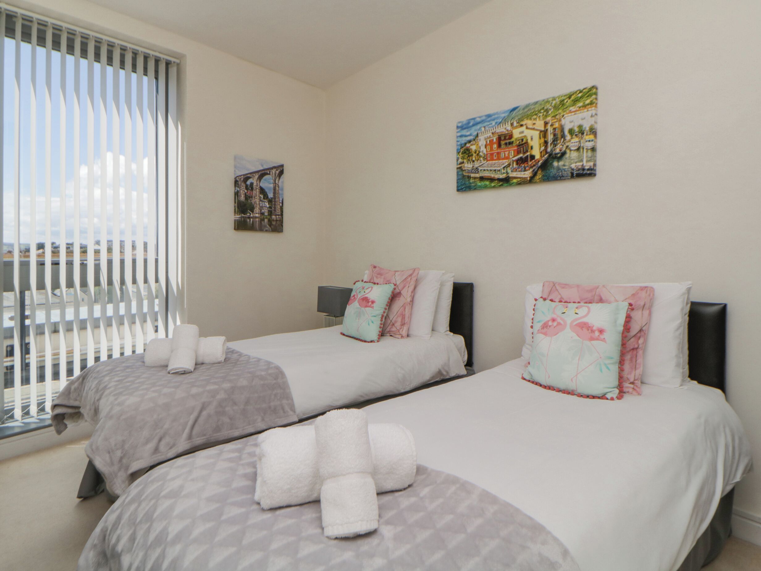A hotel room with two beds, each with a pillow and a rolled towel. There is a painting on the wall and a window with blinds.