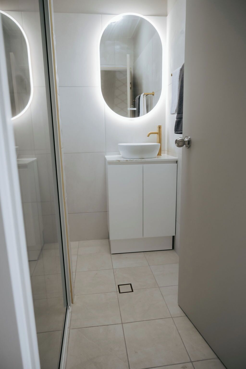 A modern bathroom with a white sink, gold faucet, and a round mirror.