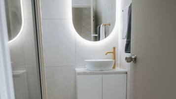 A modern bathroom with a white sink, gold faucet, and a round mirror.