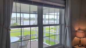 A room with a large window overlooking a grassy area and a body of water. There is a table and a chair inside the room. - Private cottage at the end of the road with hot tub, swim mat, kayaks, and a your private dock, 3 miles to the beach. (Cedar Point)