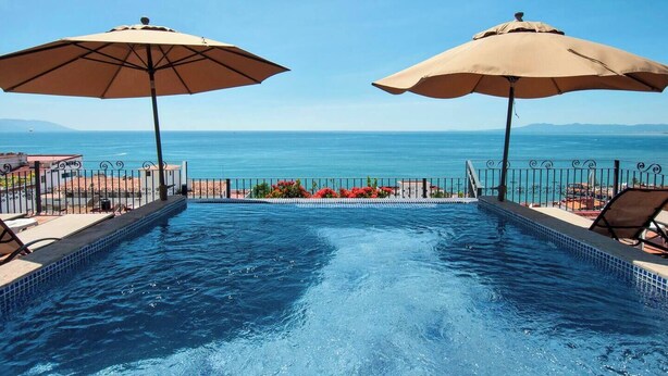 A swimming pool with two beige umbrellas, a view of the sea, and a tiled deck with a railing.