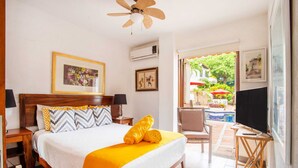 A hotel room with a bed, a ceiling fan, an air conditioning unit, a TV, and a view of an outdoor area with a pool and umbrellas.