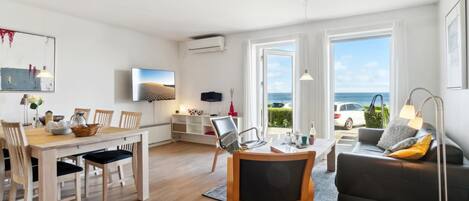 A modern living room with a dining area, a sofa, and a TV. There are large windows with a view of the sea.