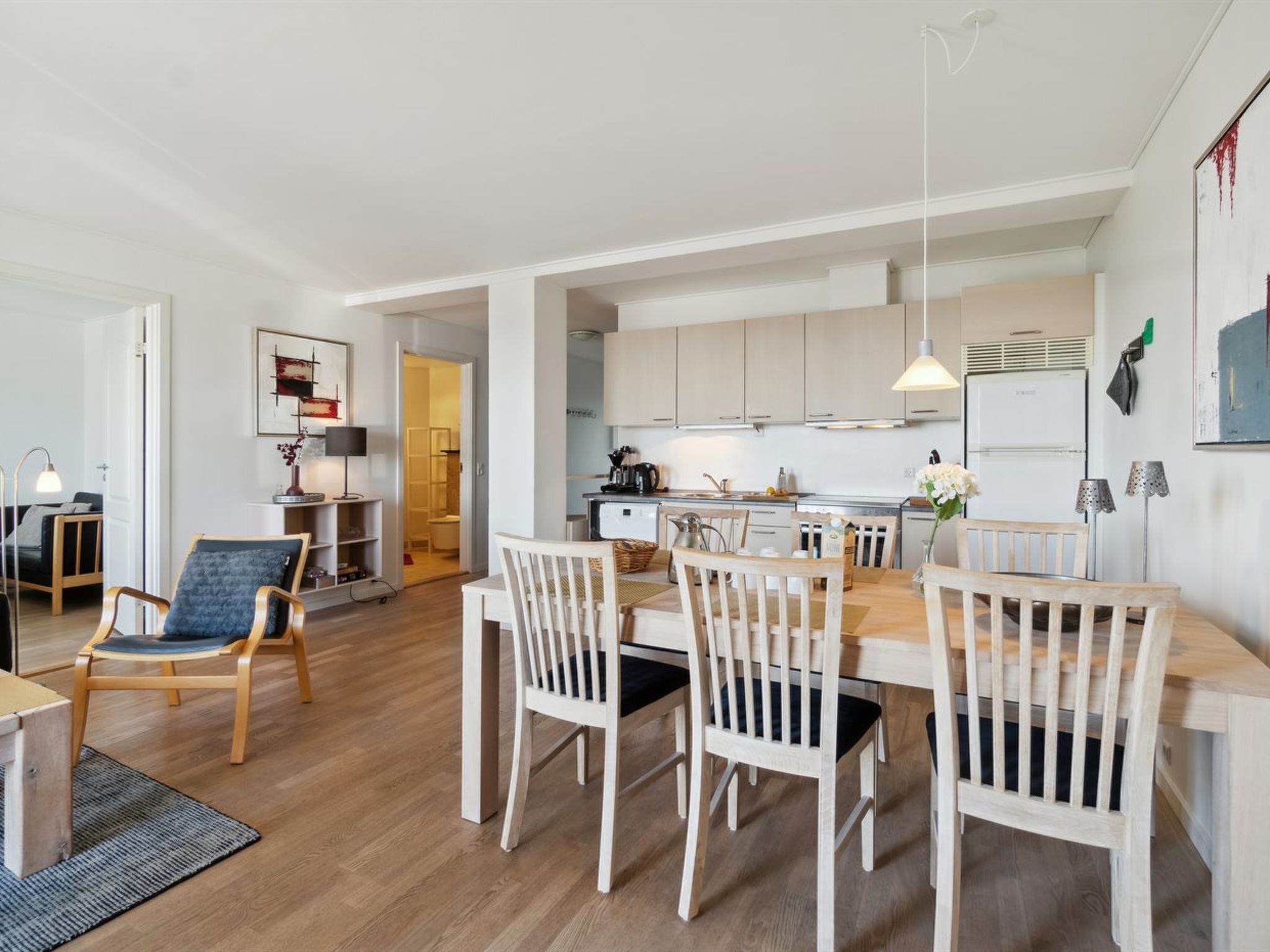 Une cuisine moderne avec des meubles blancs, un coin repas avec des chaises en bois et une salle de bain visible au fond.
