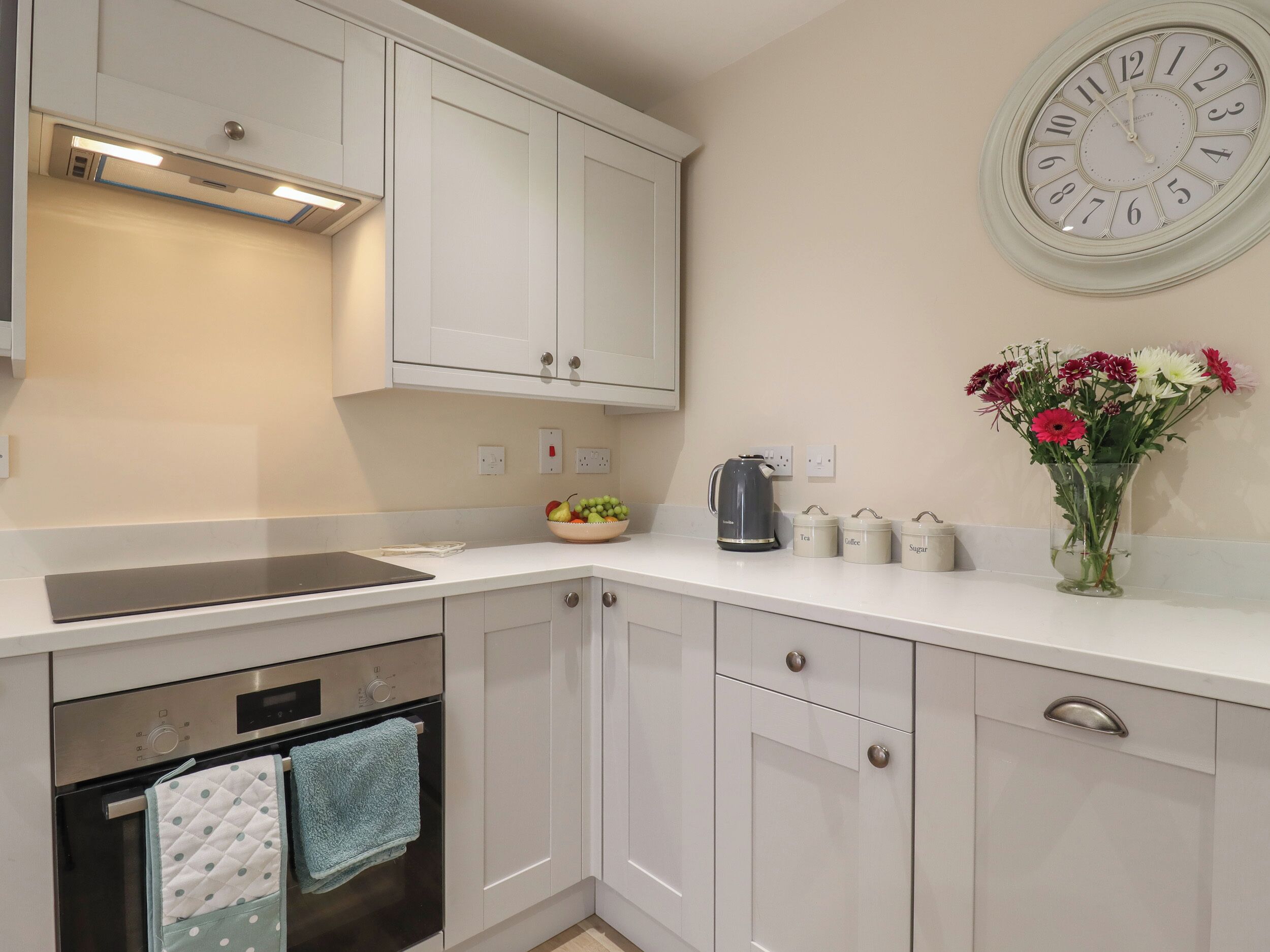 A kitchen with white cabinets, a kettle, a vase with flowers, and a clock on the wall.