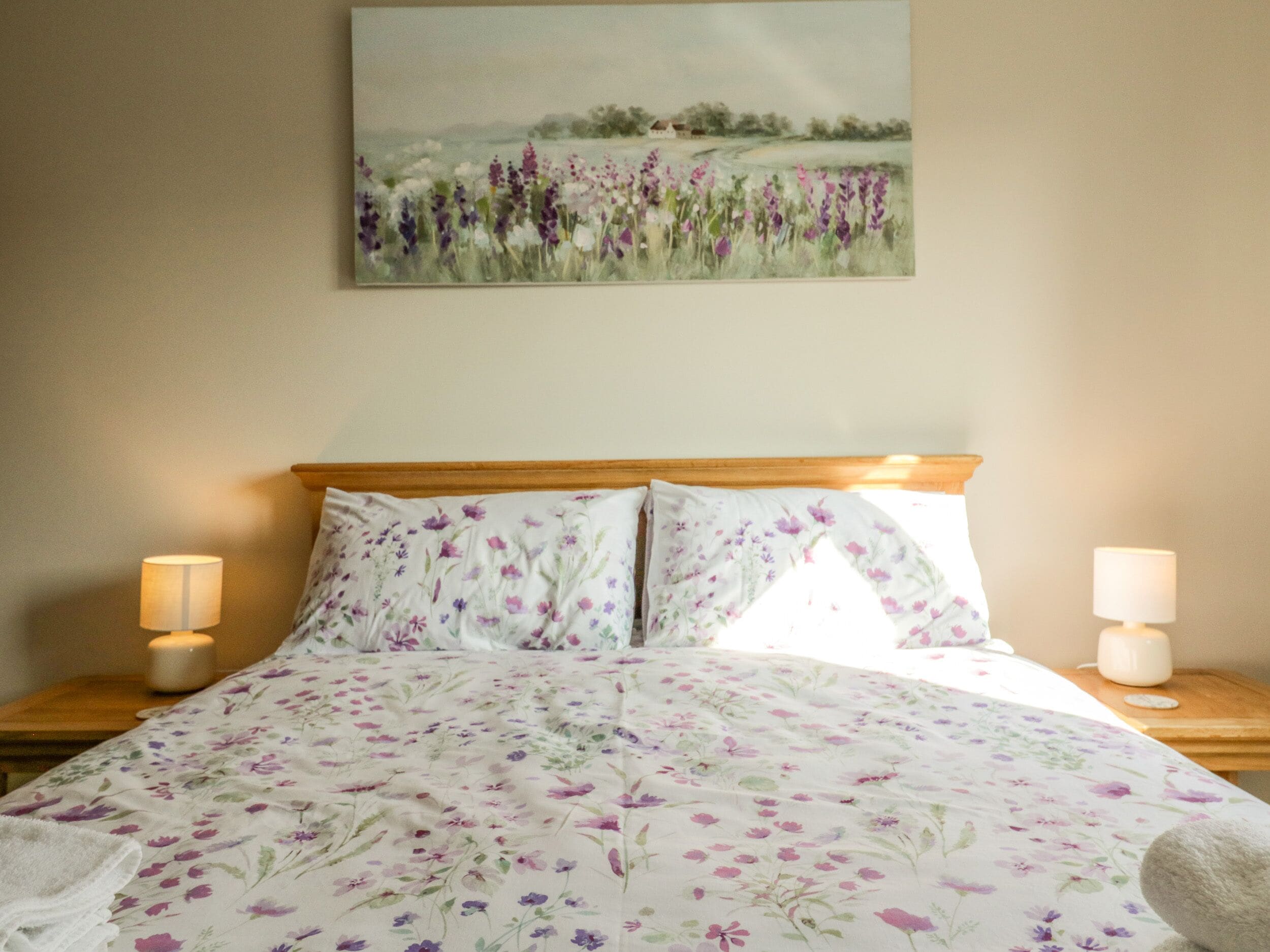 Un dormitorio con colcha de flores, dos lámparas de noche y un cuadro de un campo sobre la cama.