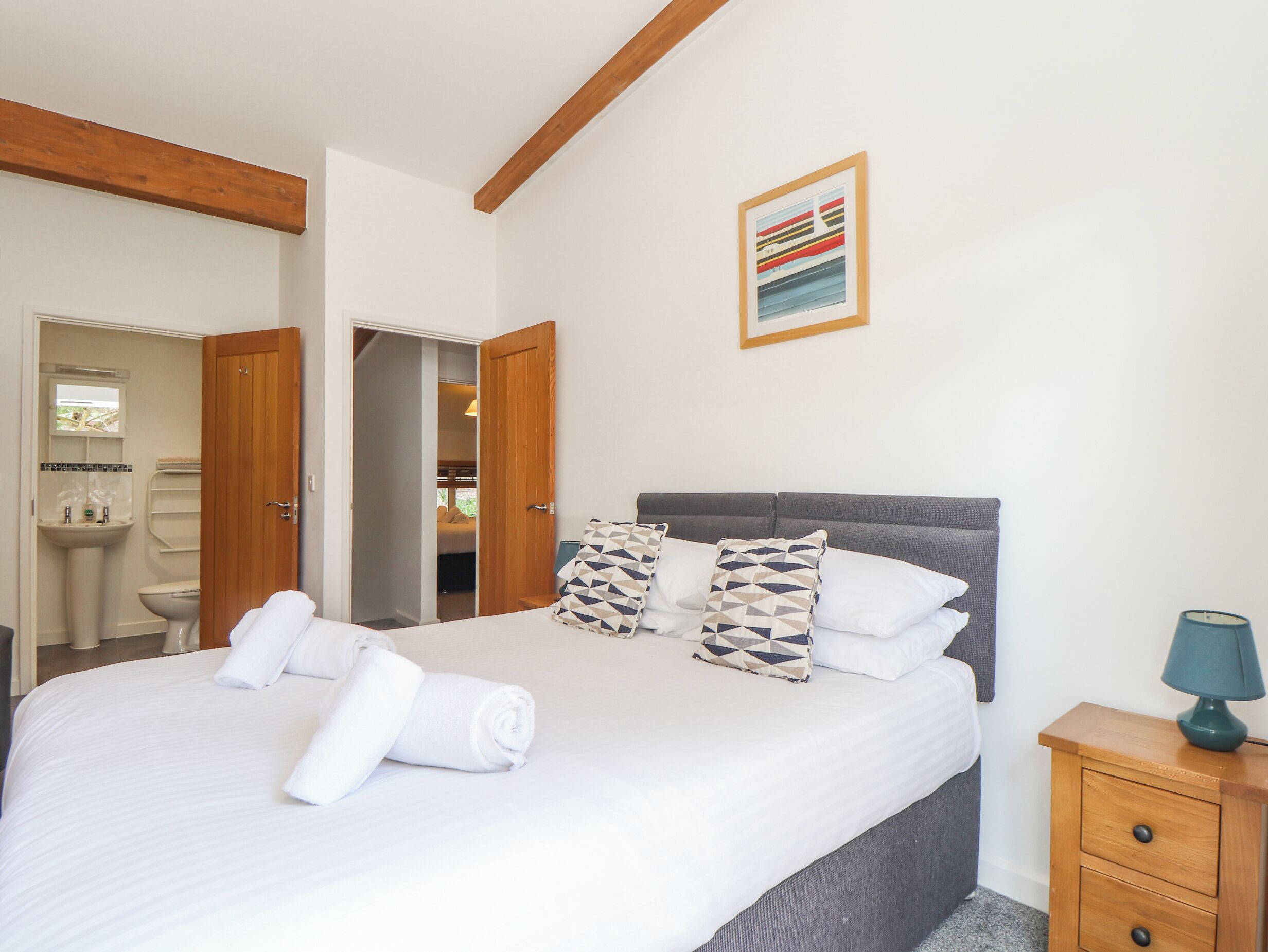 A neatly made bed with white linens and rolled towels, a gray upholstered headboard, and a wooden nightstand with a lamp.