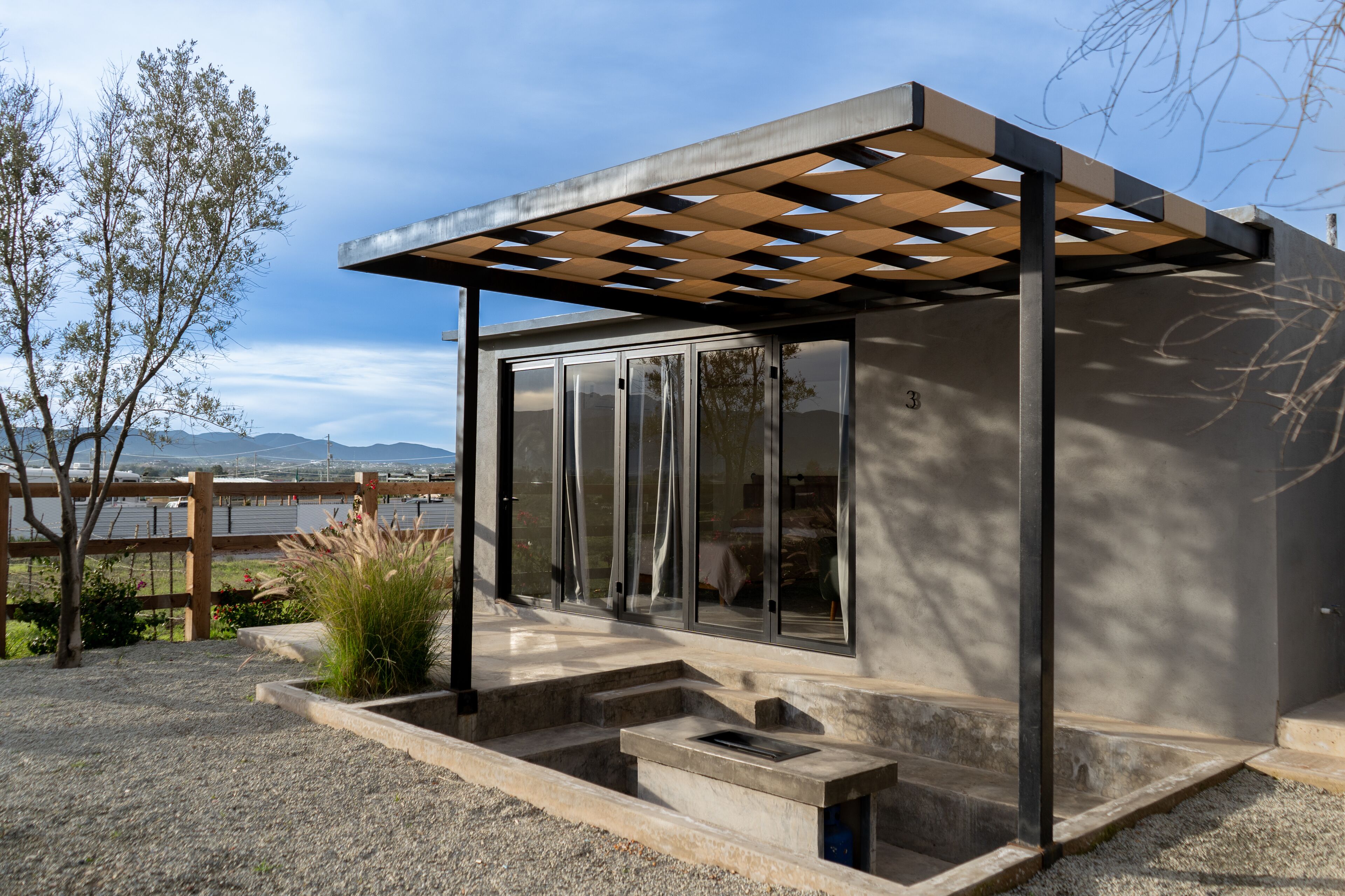 A modern outdoor structure with a pergola, steps leading to a covered area, and a view of a body of water and mountains in the background.