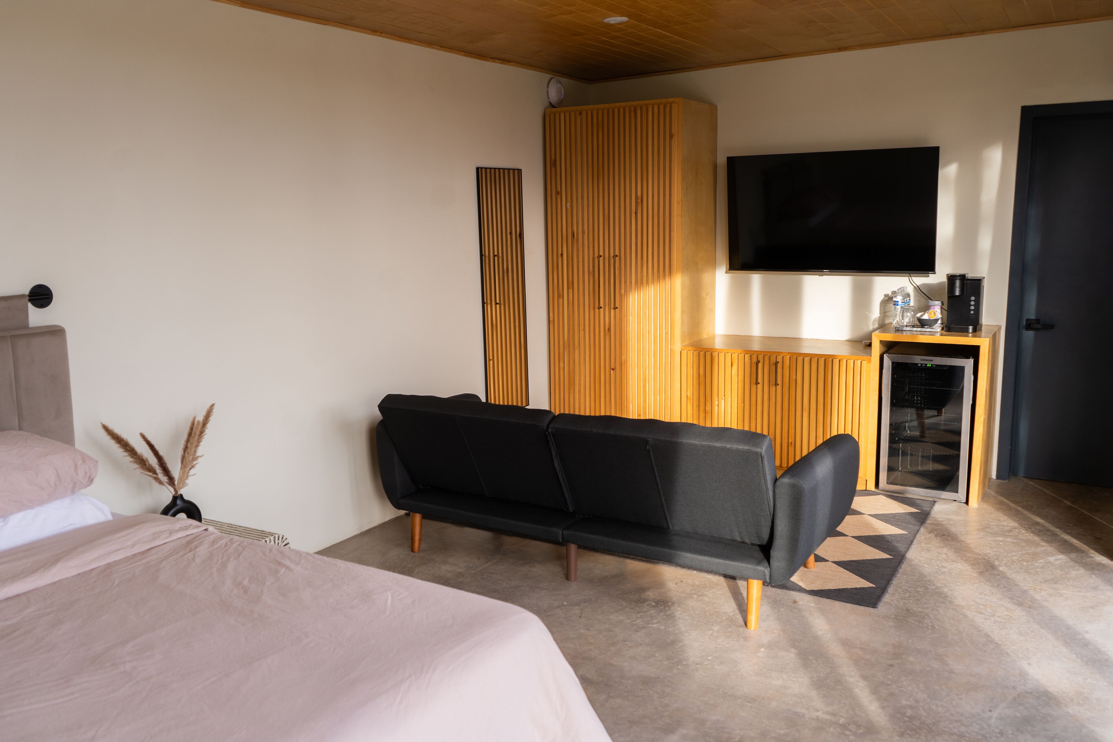 A modern bedroom with a bed, a black sofa, a wooden wardrobe, a TV, and a coffee machine.