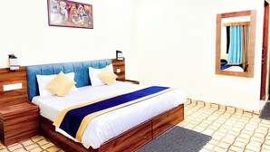 Una cama bien tendida con cabecera azul, ropa de cama blanca y una manta azul y dorada. Hay una mesita de noche de madera con una lámpara, un cuadro colgado en la pared y un espejo con cortinas.