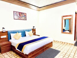 Una cama bien tendida con cabecera azul, ropa de cama blanca y una manta azul y dorada. Hay una mesita de noche de madera con una lámpara, un cuadro colgado en la pared y un espejo con cortinas.