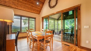 A dining area with a wooden table and chairs, large windows, and a view of greenery. - Maple Cottage in Sutton Knob (Pisgah Forest)