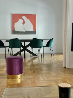 Un comedor con suelo de madera en diseño espiga, una mesa con cuatro sillas tapizadas en verde, un reposapiés morado y un cuadro abstracto enmarcado en la pared.