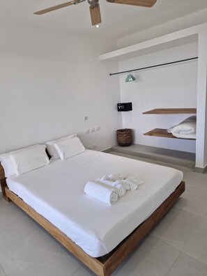A modern bedroom with a large bed, wooden headboard, a ceiling fan, a wall-mounted TV, and a built-in shelf with towels.