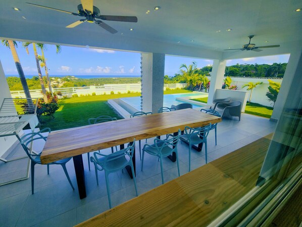 A modern outdoor dining area with a wooden table and chairs, overlooking a swimming pool and lush greenery.