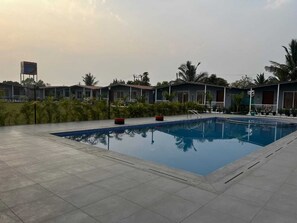 A swimming pool with a tiled deck, surrounded by small buildings and palm trees. - Omkara Resort (Rajkot)