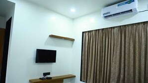 Une chambre d’hôtel équipée d’un téléviseur fixé au mur, d’une petite étagère en bois, d’une fenêtre avec des rideaux et d’un appareil de climatisation.