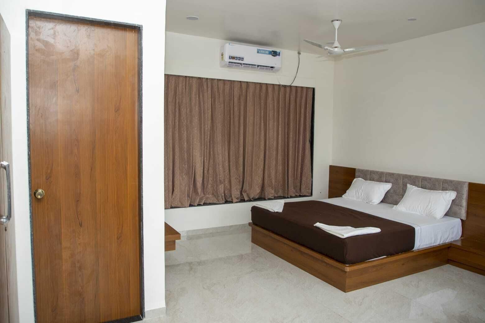 Une chambre avec une porte en bois, un ventilateur de plafond, un appareil de climatisation, un lit avec une literie blanche et marron, et une fenêtre avec des rideaux marron.