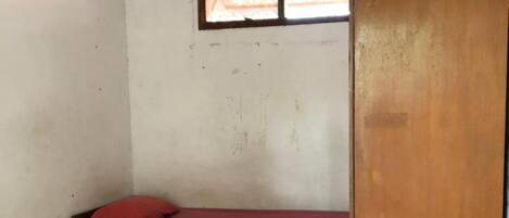 Una habitación pequeña con una cama individual, un cabecero de madera, una colcha roja y una ventana sobre la cama.