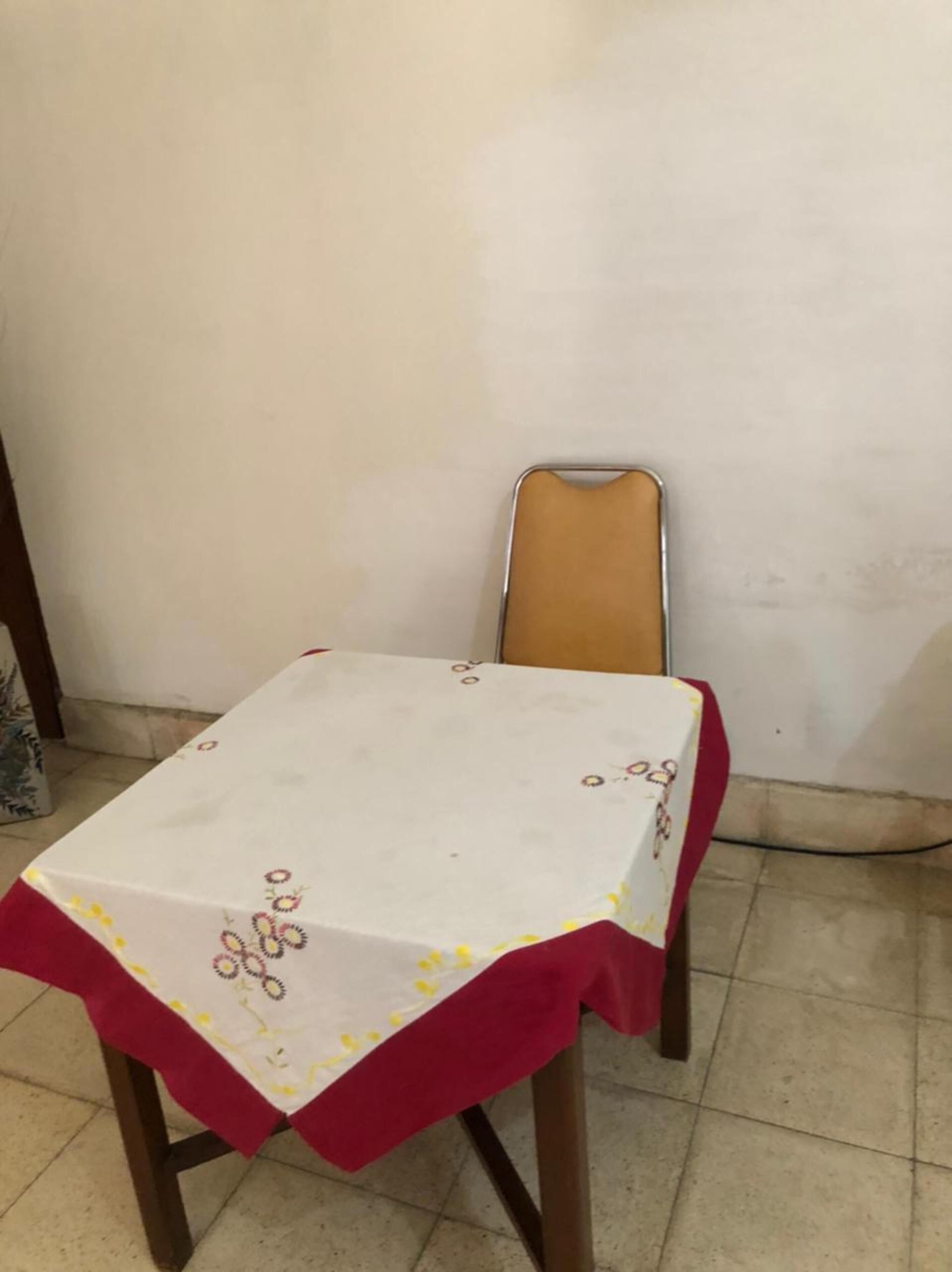 Una pequeña mesa de comedor con mantel blanco y borde rojo, junto a una única silla con estructura metálica y asiento marrón.
