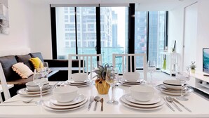 Uma sala de jantar com uma mesa posta para quatro pessoas, cadeiras brancas e vista para edifícios altos através de amplas janelas.