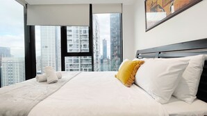 Quarto de hotel com uma cama grande, roupa de cama branca, um travesseiro amarelo e vista para edifícios altos através de janelas amplas.