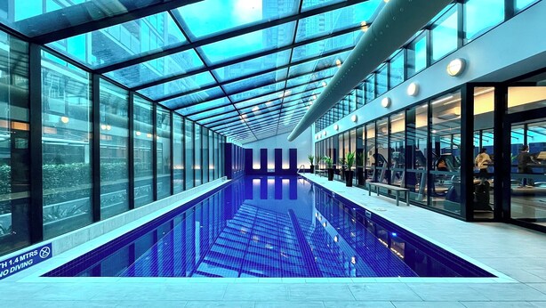 Piscina interior moderna com paredes de vidro e teto alto, rodeada por uma área comercial.