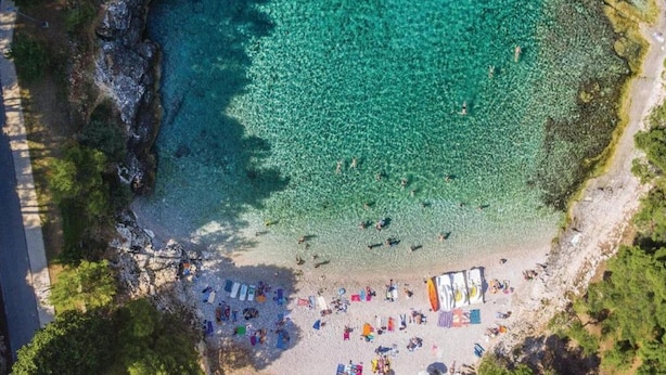 Una vista aerea di una spiaggia con acque cristalline turchesi, una riva sabbiosa e diversi ombrelloni e sdraie colorate.