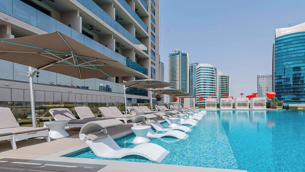 Uma área à beira da piscina com espreguiçadeiras e guarda-sóis, rodeada por edifícios modernos.